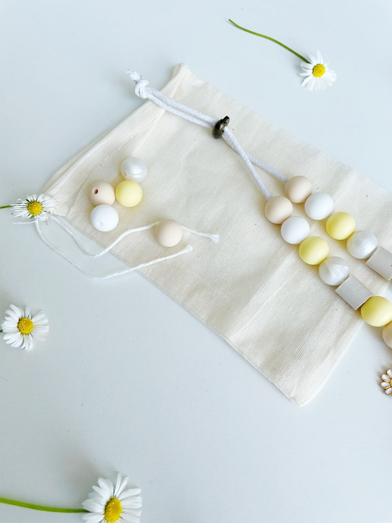 May include: A beige cotton drawstring pouch accompanied by a beaded accessory featuring round white, yellow, and tan beads, along with square grey beads. Several loose beads in matching colors are scattered near the pouch, which is decorated with white daisy flowers.