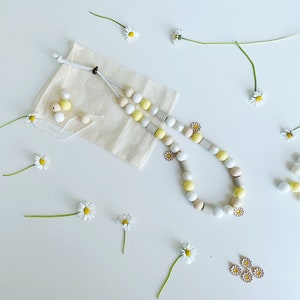 May include: A beaded necklace featuring round and cylindrical beads in shades of white, yellow, and beige, accented with small daisy-shaped charms. The necklace is accompanied by a small beige fabric drawstring pouch and loose beads.
