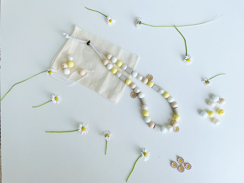 May include: A beaded necklace featuring round and cylindrical beads in shades of white, yellow, and beige, accented with small daisy-shaped charms. The necklace is accompanied by a small beige fabric drawstring pouch and loose beads.