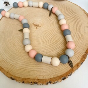May include: A necklace featuring a sequence of round and cylindrical beads in shades of grey, peach, and cream, strung on a white cord. The design includes small metallic feather-shaped charms interspersed among the beads, creating a textured look.
