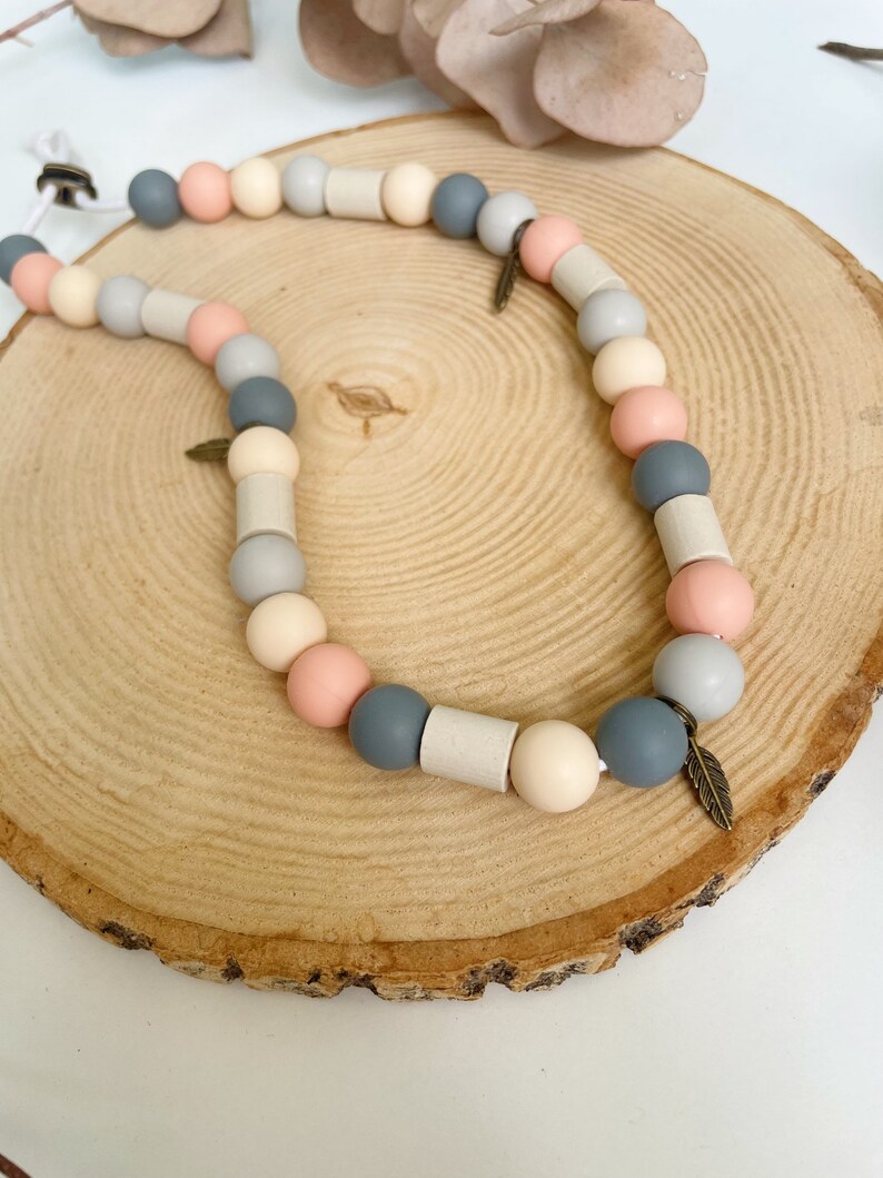 May include: A necklace featuring a sequence of round and cylindrical beads in shades of grey, peach, and cream, strung on a white cord. The design includes small metallic feather-shaped charms interspersed among the beads, creating a textured look.