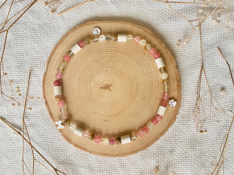 May include: A circular beaded necklace featuring translucent pink and yellow beads, interspersed with cylindrical white spacers. The necklace includes a small circular charm with the letter L, a white bone charm, and a white paw print charm.