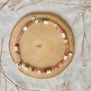 May include: A beaded bracelet featuring translucent pink and yellow beads, interspersed with cylindrical white spacers. The bracelet includes a small white circular charm with the letter L, a white paw print charm, and a small white bone-shaped charm.