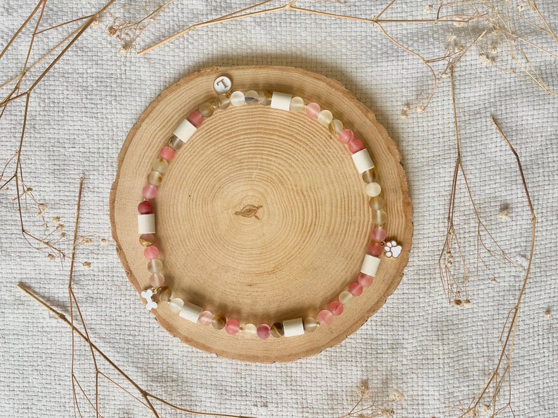 May include: A beaded bracelet featuring translucent pink and yellow beads, interspersed with cylindrical white spacers. The bracelet includes a small white circular charm with the letter L, a white paw print charm, and a small white bone-shaped charm.