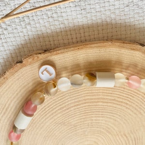 May include: A beaded necklace featuring a mix of translucent pink, beige, and brown frosted beads interspersed with smooth white cylindrical beads. A circular white charm with a gold-colored letter L is attached to the strand of beads.