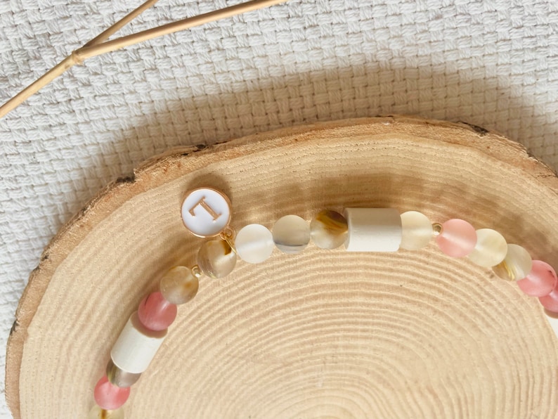 May include: A beaded necklace featuring a mix of translucent pink, beige, and brown frosted beads interspersed with smooth white cylindrical beads. A circular white charm with a gold-colored letter L is attached to the strand of beads.