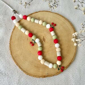 May include: A beaded lanyard featuring a repeating pattern of round and cylindrical beads in shades of red, sage green, white, and cream. The accessory is accented with three small, dangling cherry charms, each featuring two red enamel cherries with green leaves.