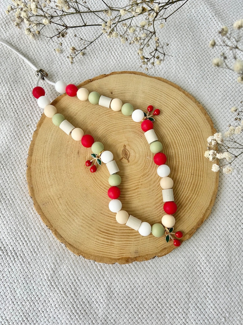 May include: A beaded lanyard featuring a repeating pattern of round and cylindrical beads in shades of red, sage green, white, and cream. The accessory is accented with three small, dangling cherry charms, each featuring two red enamel cherries with green leaves.
