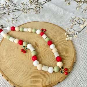 May include: A beaded necklace featuring a mix of round and cylindrical beads in shades of red, white, cream, and sage green. The necklace is accented with small gold-toned metal charms shaped like pairs of cherries with green enamel leaves.