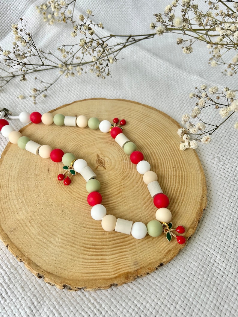 May include: A beaded necklace featuring a mix of round and cylindrical beads in shades of red, white, cream, and sage green. The necklace is accented with small gold-toned metal charms shaped like pairs of cherries with green enamel leaves.