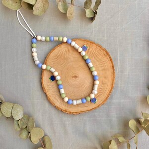 May include: A phone charm featuring a white cord loop attached to a strand of beads in shades of light blue, sage green, white, and cream. The strand includes cylindrical and spherical beads, accented by two small blue berry-shaped charms.