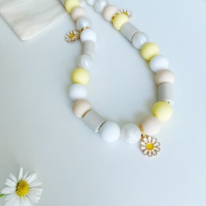 May include: A beaded necklace featuring a repeating pattern of round and cylindrical beads in shades of white, cream, pale yellow, and light gray. The necklace is accented with small, gold-toned metal daisy charms with white and yellow enamel petals.
