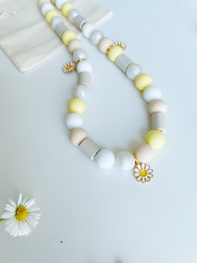 May include: A beaded necklace featuring a repeating pattern of round and cylindrical beads in shades of white, cream, pale yellow, and light gray. The necklace is accented with small, gold-toned metal daisy charms with white and yellow enamel petals.