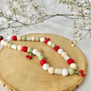 May include: A beaded necklace featuring a repeating pattern of round and cylindrical beads in shades of red, sage green, cream, and white. The necklace is accented with two small gold-toned metal charms shaped like pairs of red cherries with green leaves.