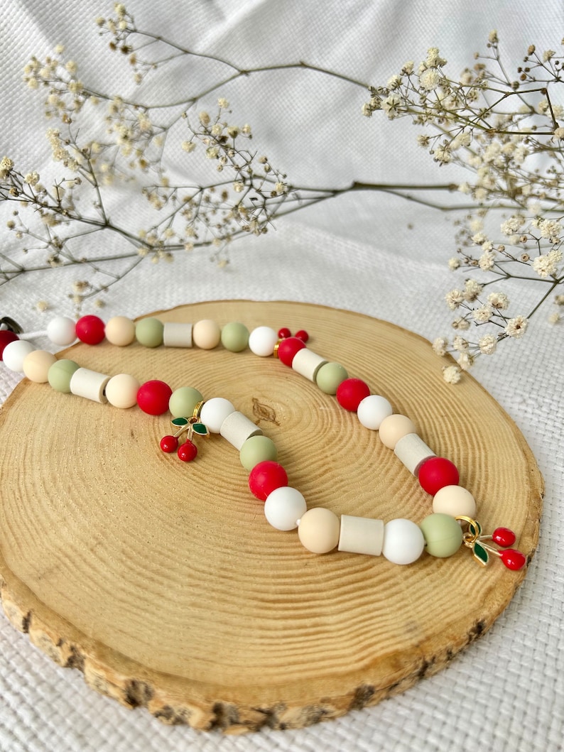 May include: A beaded necklace featuring a repeating pattern of round and cylindrical beads in shades of red, sage green, cream, and white. The necklace is accented with two small gold-toned metal charms shaped like pairs of red cherries with green leaves.