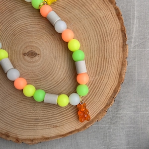 May include: A beaded necklace featuring a repeating pattern of neon yellow, lime green, orange, and white spherical beads, interspersed with cylindrical grey beads. The necklace is accented with two translucent gummy bear charms.