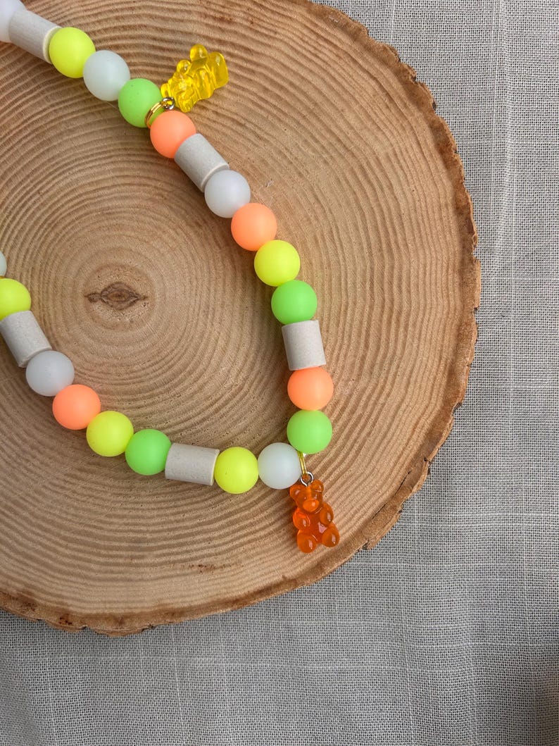 May include: A beaded necklace featuring a repeating pattern of neon yellow, lime green, orange, and white spherical beads, interspersed with cylindrical grey beads. The necklace is accented with two translucent gummy bear charms.