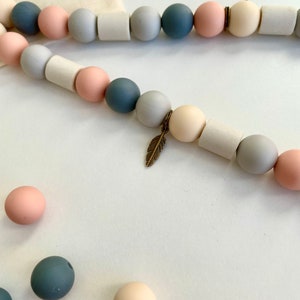 May include: A string of beads featuring alternating spherical and cylindrical shapes in shades of dusty blue, soft pink, and cream. The strand includes a small, metallic bronze-coloured feather charm suspended between two beads, with loose beads nearby.