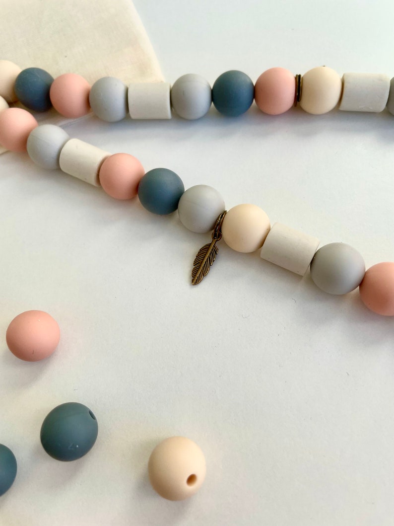 May include: A string of beads featuring alternating spherical and cylindrical shapes in shades of dusty blue, soft pink, and cream. The strand includes a small, metallic bronze-coloured feather charm suspended between two beads, with loose beads nearby.