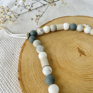 May include: A beaded accessory featuring a sequence of round and cylindrical beads in shades of white, light gray, dark gray, and marble patterns. The strand includes a white cord and a small white bone-shaped charm attached with a gold-toned ring.