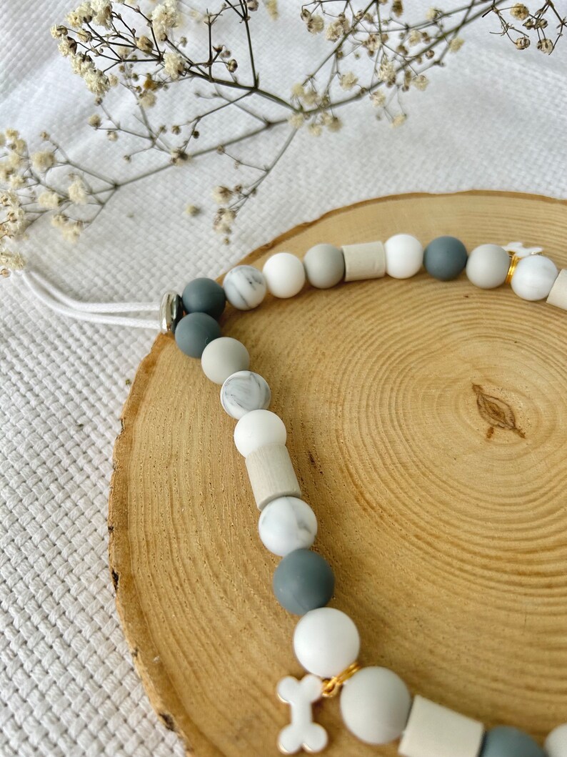May include: A beaded accessory featuring a sequence of round and cylindrical beads in shades of white, light gray, dark gray, and marble patterns. The strand includes a white cord and a small white bone-shaped charm attached with a gold-toned ring.