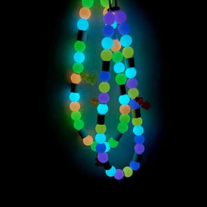 May include: A glowing beaded necklace featuring round beads in shades of neon green, blue, orange, and purple, interspersed with small cylindrical spacers and tiny bear-shaped charms. The necklace emits a bright light against a dark background.