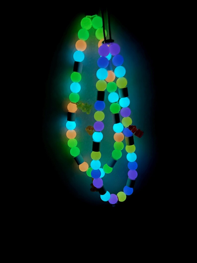 May include: A glowing beaded necklace featuring round beads in shades of neon green, blue, orange, and purple, interspersed with small cylindrical spacers and tiny bear-shaped charms. The necklace emits a bright light against a dark background.