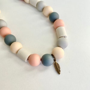 May include: A necklace featuring a repeating pattern of round and cylindrical beads in shades of dusty pink, slate blue, cream, and light grey. The strand includes a small, metallic bronze-toned feather charm attached to one of the beads.
