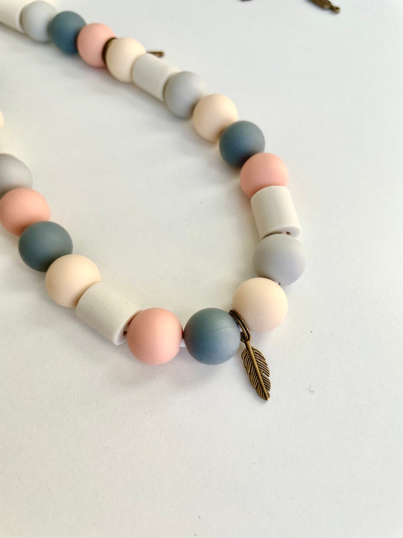 May include: A necklace featuring a repeating pattern of round and cylindrical beads in shades of dusty pink, slate blue, cream, and light grey. The strand includes a small, metallic bronze-toned feather charm attached to one of the beads.