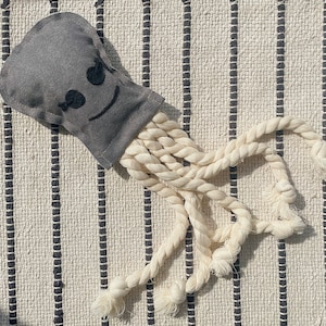 May include: A gray fabric octopus toy with a smiling face and white rope tentacles. The toy is lying on a white and black striped surface.