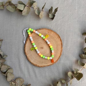 May include: A beaded necklace featuring a repeating pattern of round neon green, orange, and white beads. The necklace is accented with three small, translucent gummy bear-shaped charms in yellow, green, and orange, strung on a white cord.