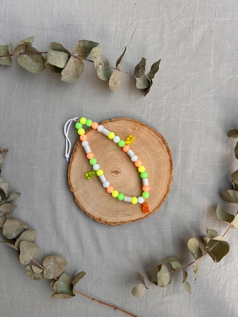 May include: A beaded necklace featuring a repeating pattern of round neon green, orange, and white beads. The necklace is accented with three small, translucent gummy bear-shaped charms in yellow, green, and orange, strung on a white cord.