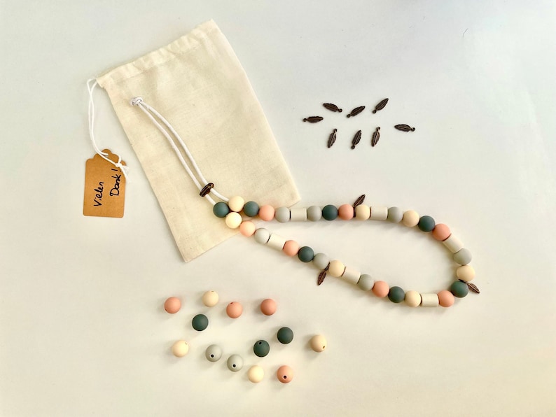 May include: A long necklace made of round and cylindrical silicone beads in shades of sage green, dusty pink, cream, and grey, featuring small dark brown leaf-shaped charms. Includes a beige fabric drawstring pouch and a tag reading Vielen Dank.