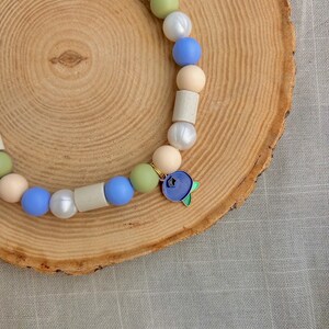 May include: A beaded necklace featuring a sequence of round and cylindrical beads in shades of blue, sage green, white, and beige. The necklace includes a single metal charm shaped like a blueberry with green leaves and a small star cutout.