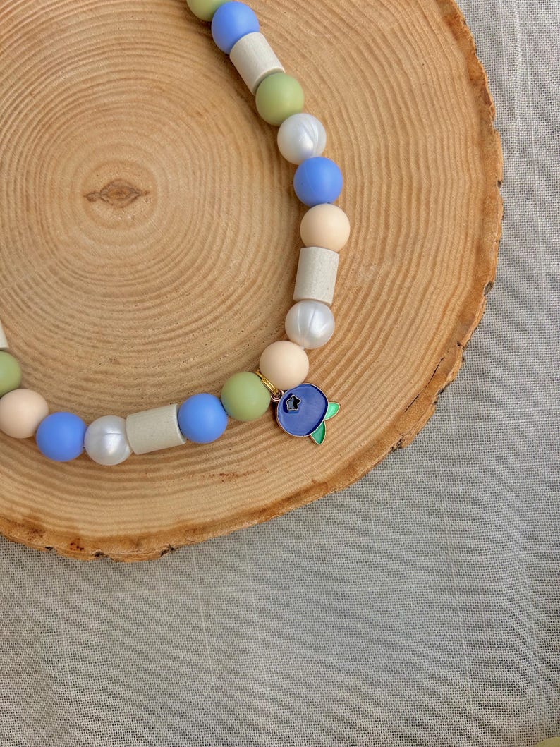 May include: A beaded necklace featuring a sequence of round and cylindrical beads in shades of blue, sage green, white, and beige. The necklace includes a single metal charm shaped like a blueberry with green leaves and a small star cutout.