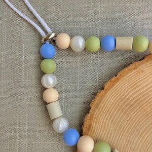 May include: A beaded lanyard featuring a white cord and a silver-toned metal connector. The strand consists of alternating round and cylindrical beads in matte and pearlescent finishes, colored in shades of light blue, sage green, cream, and white.