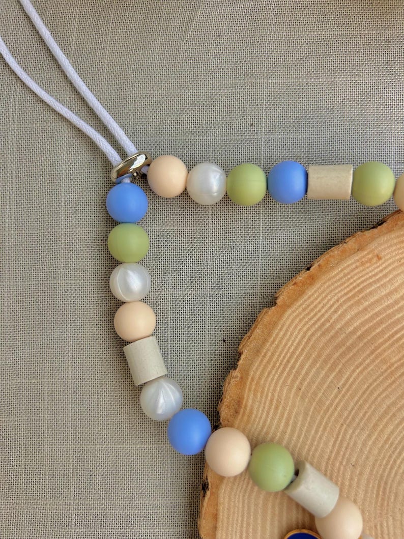 May include: A beaded lanyard featuring a white cord and a silver-toned metal connector. The strand consists of alternating round and cylindrical beads in matte and pearlescent finishes, colored in shades of light blue, sage green, cream, and white.