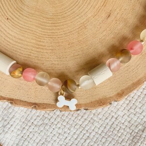 May include: A beaded necklace featuring a central white enamel dog bone charm. The necklace is composed of alternating matte and translucent beads in shades of pink, cream, and brown, interspersed with cylindrical off-white wooden beads.
