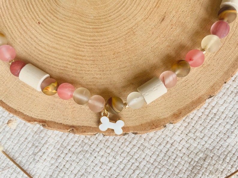 May include: A beaded necklace featuring a central white enamel dog bone charm. The necklace is composed of alternating matte and translucent beads in shades of pink, cream, and brown, interspersed with cylindrical off-white wooden beads.