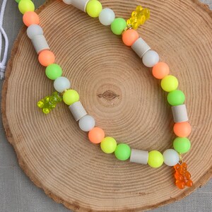 May include: A beaded lanyard featuring a repeating pattern of round neon green, bright orange, and matte white cylindrical beads. Three translucent gummy bear charms in yellow, lime green, and orange are attached to the strand with gold jump rings.