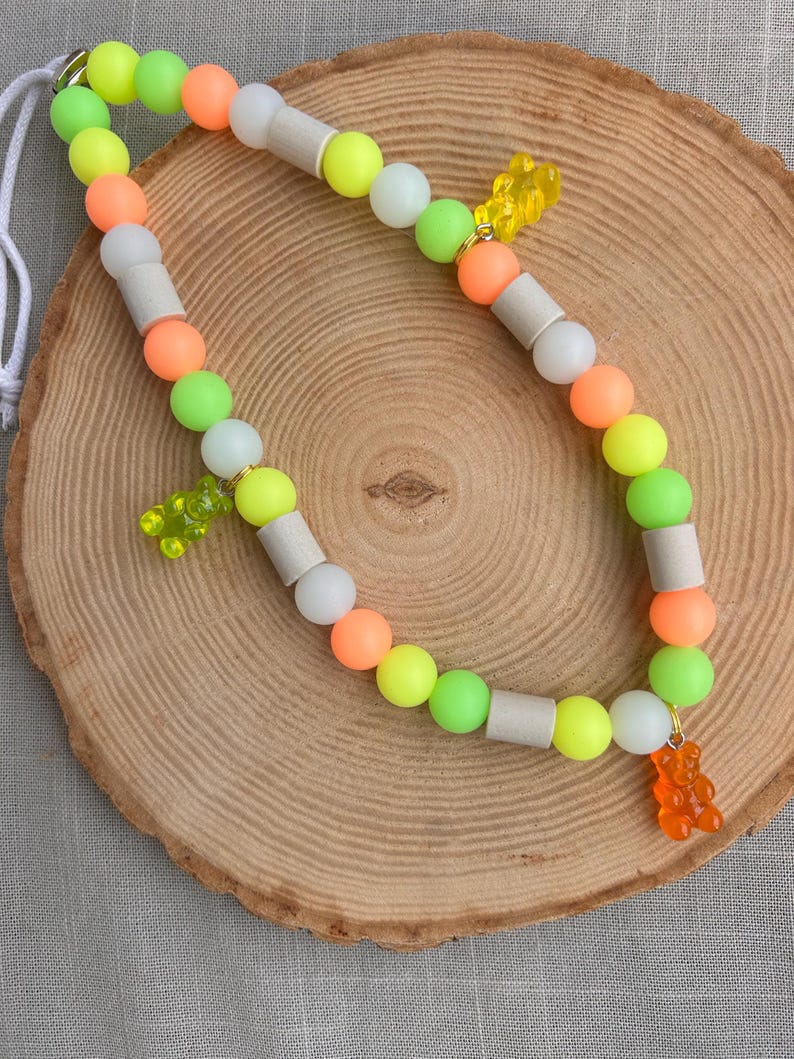 May include: A beaded lanyard featuring a repeating pattern of round neon green, bright orange, and matte white cylindrical beads. Three translucent gummy bear charms in yellow, lime green, and orange are attached to the strand with gold jump rings.