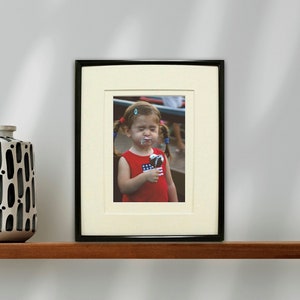 May include: A framed photograph of a young girl with pigtails eating an ice cream cone. She is wearing a red shirt with white stars and a white headband. The frame is black with a white mat.
