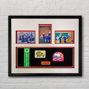 May include: A black framed shadow box with a white mat and red borders. The shadow box contains four photos of a group of people, a green ribbon with the word "Participant" on it, and three patches. The patches are a yellow truck with the words "Postcards for Food" on it, a red, white, and blue wagon with the words "Don't Disconnect from Your Roots" on it, and a red and white patch with the words "Tiger Cubs BSA" on it.