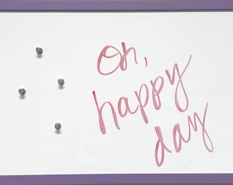 May include: A white dry erase board with a purple frame. The board has three silver push pins and the words "Oh, happy day" written in pink marker.