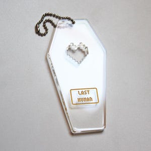 以下が含まれることがあります： ピクセル化されたハートの切り抜きと「LAST HUMAN」と書かれた金色のフレームのラベルが付いた、透明な棺型のキーホルダー。 キーホルダーは、小さな濃い色の金属ボールチェーンに取り付けられています。