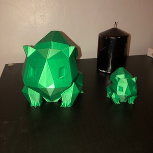 May include: Two green 3D printed figurines of a Pokemon character. The larger figurine is standing on a black surface. The smaller figurine is sitting on the surface.