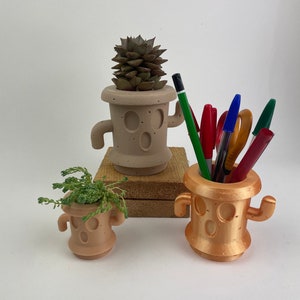 Peut inclure: Trois pots de fleurs imprimés en 3D en forme de personnages de dessins animés avec des bras et des jambes. Les pots sont beige et or. Le plus grand pot beige contient une plante succulente. Le plus petit pot beige contient une petite plante verte. Le pot doré est vide et contient un stylo vert, rouge et bleu.