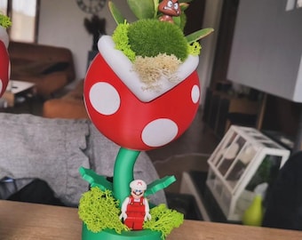 Puede incluir: Una maceta roja y blanca con forma de Planta Piraña de la serie de videojuegos Super Mario. La maceta tiene una base verde y una pequeña planta verde que crece desde la parte superior. Una pequeña figura de Mario está de pie en la maceta.