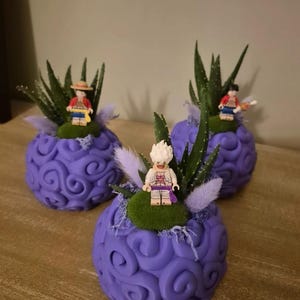 Peut inclure: Trois sculptures violettes décoratives en forme de fruit stylisé, chacune surmontée d'une figurine Lego, de mousse verte et de plantes vertes épineuses. Les sculptures sont sur une surface en bois.
