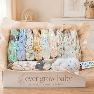 以下が含まれることがあります： ever grow babyと書かれた白い段ボール箱の中に、様々なパステルカラーの柄の折り畳まれたベビー用おむつが透明なプラスチックに包まれて入っており、carter's child of mineというタグが付いた白いぬいぐるみも添えられています。